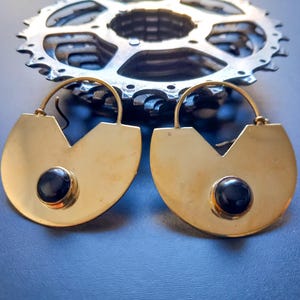 May include: A pair of gold-colored earrings with a geometric design, featuring a black stone in the center of each earring. A silver gear is in the background. The earrings have a unique, modern style.