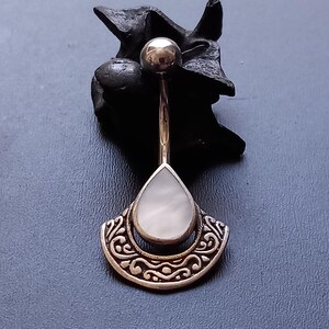Peut inclure: Un piercing de nombril argenté avec une pierre blanche en forme de larme et un motif argenté décoratif.