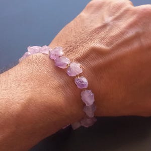 Peut inclure: Un bracelet composé de pierres d'améthyste violet clair taillées à l'état brut. Les pierres sont enfilées avec de petites perles dorées. Le bracelet est porté au poignet, sur fond bleu.
