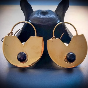 May include: A pair of gold-colored earrings with a unique design. Each earring features a rounded shape with a V-shaped cutout and a dark, round stone in the center. The earrings have a simple, circular hook.