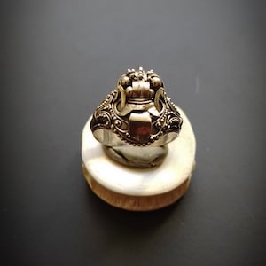 May include: A silver ring with an ornate design featuring a crown-like top and intricate details. The ring is displayed on a white stand against a grey background.