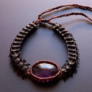 May include: A purple amethyst gemstone is woven into a brown cord bracelet. The bracelet is made of black rubber tire pieces that are shaped like a spine.