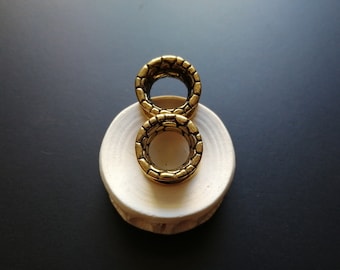 以下が含まれることがあります： 2つの金色の金属製のイヤーゲージが、白い円形のスタンドの上に置かれています。ゲージは、テクスチャ加工されたパターンデザインになっています。