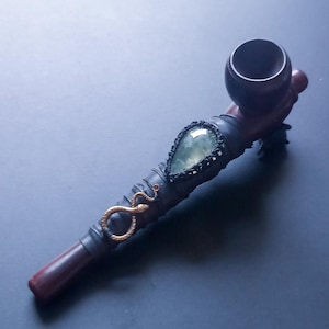 May include: A dark brown wooden smoking pipe featuring a teardrop-shaped green gemstone set in black macrame cord. The stem is wrapped in black material and adorned with a small, metallic gold-coloured snake figure attached to the side.