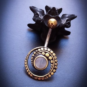 Puede incluir: Un piercing para el ombligo con un diseño en espiral dorado y una piedra central de color claro. El anillo tiene una bola dorada en la parte superior, sobre un fondo oscuro.