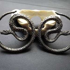 May include: A pair of silver hoop earrings featuring a snake design. The snakes are coiled around the hoops and have a textured surface.