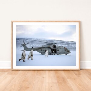 May include: A framed print featuring a gray helicopter on a snowy landscape. Three figures in white camouflage are near the helicopter. The background shows snow-covered mountains and a cloudy sky. The print is in a light brown wooden frame.