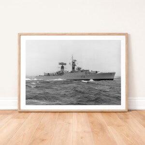 Op de afbeelding: Zwart-witfoto van een oorlogsschip op zee. Het schip vaart door ruw water, met golven zichtbaar rond de romp. Het schip heeft een hoge mast en diverse antennes en radarapparatuur. De scheepsromp is gemarkeerd met het nummer "F14".