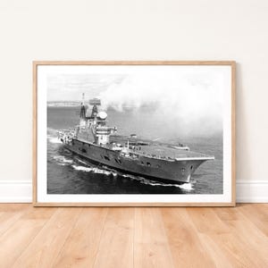 May include: A monochrome photograph of a naval aircraft carrier at sea. The ship is moving through the water, with a large flight deck and a prominent superstructure. A plume of steam rises in the background.