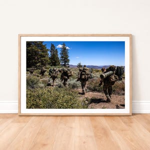 May include: A framed print depicts a group of people in military attire hiking through a natural landscape. They carry large packs and equipment, walking on a dirt path under a clear blue sky. The scene is set amidst green foliage and tall trees.