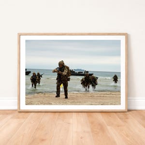 May include: A framed print depicts soldiers in camouflage uniforms wading through water onto a beach. They are carrying rifles and appear to be part of a military exercise. The sky is overcast, and boats are visible in the background.