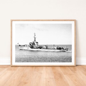 Peut inclure: Tirage encadré en noir et blanc d'un navire de guerre en mer. Le bateau, avec un mât imposant et des structures de pont, est en mouvement. L'impression est posée sur un sol en bois clair.