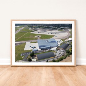 Può includere: Vista aerea di un aeroporto con diversi aerei sulla pista e in volo. L'immagine mostra un grande hangar, piste e campi verdi circostanti. La fotografia è incorniciata in una cornice di legno chiaro.