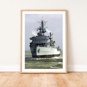 May include: A framed print showing a grey warship at sea. The ship has a tall superstructure with masts and antennas. The hull is black. The sea is green and choppy. The print is in a light wood frame.