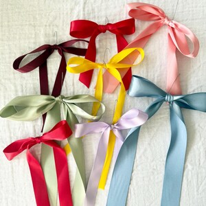 Puede incluir: Una colección de lazos de cinta de raso de colores, en tonos como rojo, amarillo, rosa, azul y verde. Cada lazo está sujeto a un alambre fino. Los lazos están dispuestos sobre una superficie blanca.