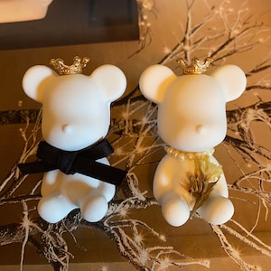 May include: Two white ceramic teddy bears wearing gold crowns. One bear has a black bow tie and the other has a pearl necklace and a dried flower.
