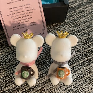 Puede incluir: Dos figuritas de oso de cerámica blanca con coronas doradas, cada una sosteniendo una taza de cerámica marrón con un logotipo de Starbucks verde. Los osos llevan bufandas rosa y gris. Los osos están sentados sobre una superficie gris texturizada.