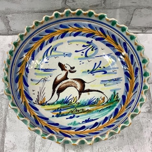 May include: A hand-painted ceramic plate with a scalloped edge. The plate features a brown dog in a field, surrounded by blue, yellow, and green decorative elements. The rim has a blue and green design.