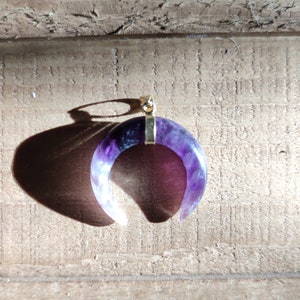 K&ouml;nnte beinhalten: Ein Halbmond-Anh&auml;nger aus Amethyst mit einer goldfarbenen &Ouml;se. Der Amethyst ist tiefviolett mit wei&szlig;en Streifen.