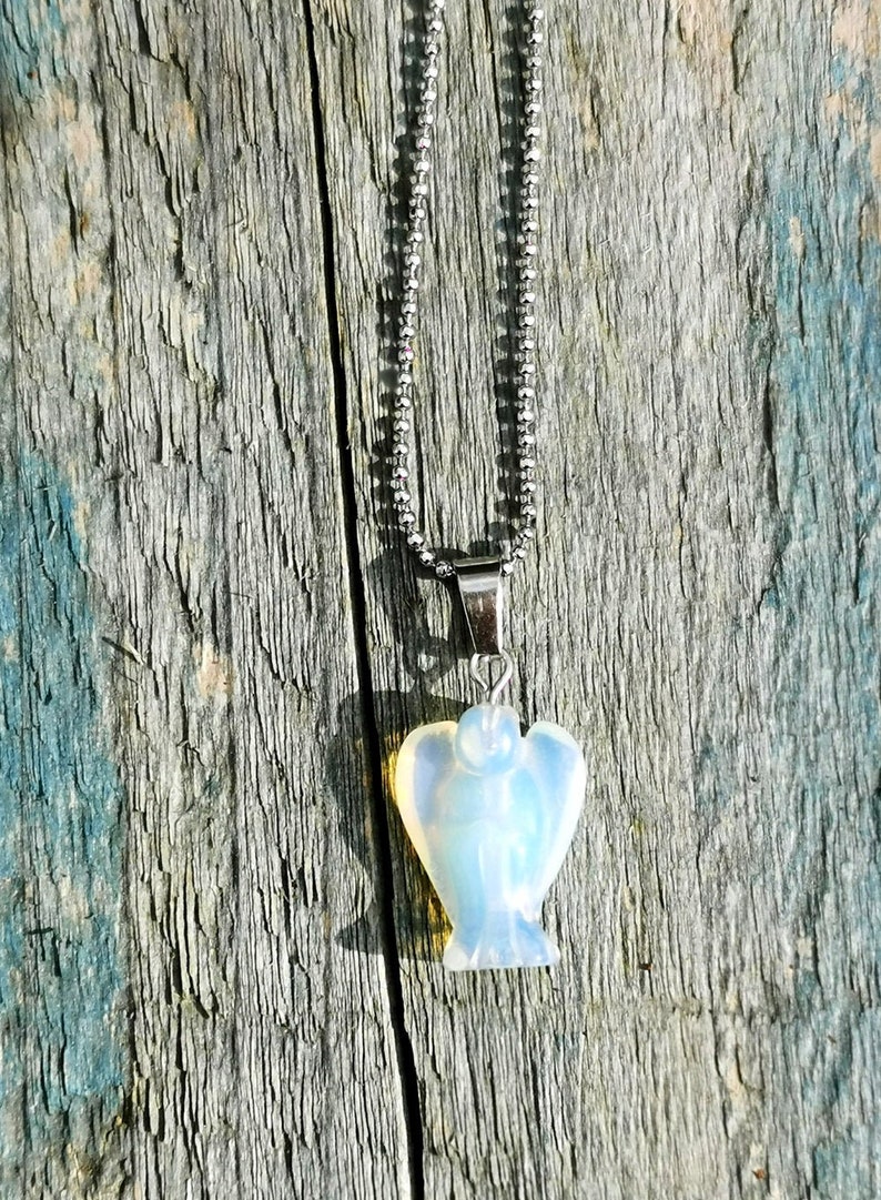 Op de afbeelding: Een zilveren ketting met een witte opaal engel hanger. De hanger is gegraveerd in de vorm van een engel met uitgespreide vleugels. De ketting is gefotografeerd tegen een verweerde houten achtergrond.