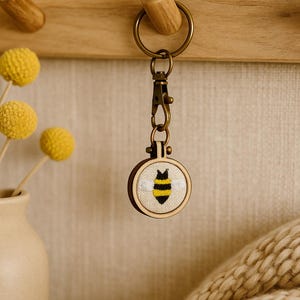 May include: A round wooden keyring featuring a stitched bee design. The bee is yellow and black with white wings, set against a beige background. The keyring has a bronze-coloured clasp and ring.