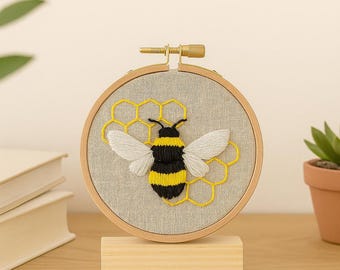 May include: Embroidered bee artwork in a wooden hoop. The bee is black and yellow with white wings, set against a grey fabric background. A yellow honeycomb design surrounds the bee. The artwork is displayed on a wooden stand.