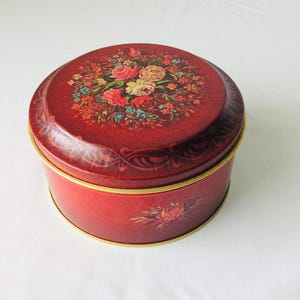 May include: A vintage red tin with a floral design on the lid and a rose motif on the side. The tin has a gold-coloured rim and a crackled finish, giving it a rustic appearance. The floral arrangement features roses and other flowers.