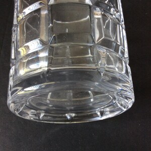 Op de afbeelding: Een glazen vat van helder glas met een geometrisch rasterpatroon van verhoogde vierkanten over het cilindrische lichaam. De basis is cirkelvormig met een dikke, gladde rand en het glasoppervlak reflecteert het licht, wat de gefacetteerde textuur benadrukt.
