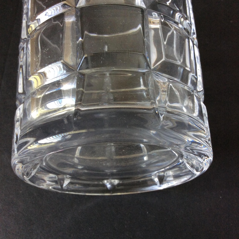 Op de afbeelding: Een glazen vat van helder glas met een geometrisch rasterpatroon van verhoogde vierkanten over het cilindrische lichaam. De basis is cirkelvormig met een dikke, gladde rand en het glasoppervlak reflecteert het licht, wat de gefacetteerde textuur benadrukt.