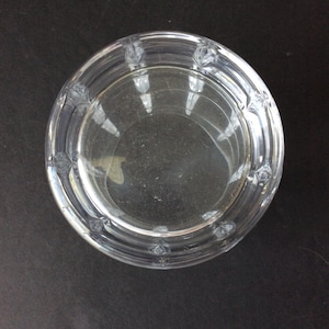 Op de afbeelding: Een ronde kom van helder glas met een gladde binnenkant en een dikke, getextureerde buitenrand. De rand is versierd met een herhalend patroon van kleine, verhoogde geometrische inkepingen die zorgen voor een gefacetteerd uiterlijk rondom.