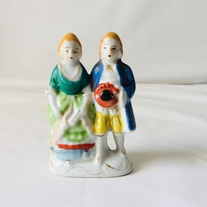 May include: A small porcelain figurine depicting a couple in historical attire. The woman wears a green dress with a white apron, while the man is in a blue coat and yellow breeches. The figurine is painted with bright colors and has gold accents.