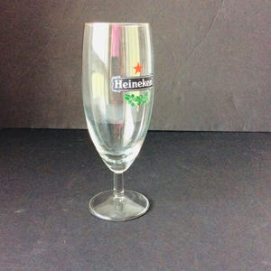 May include: A clear, stemmed beer glass with a wide bowl and a narrow base. The glass features the Heineken logo, including a red star and green accents. The glass is empty and sits on a dark surface.