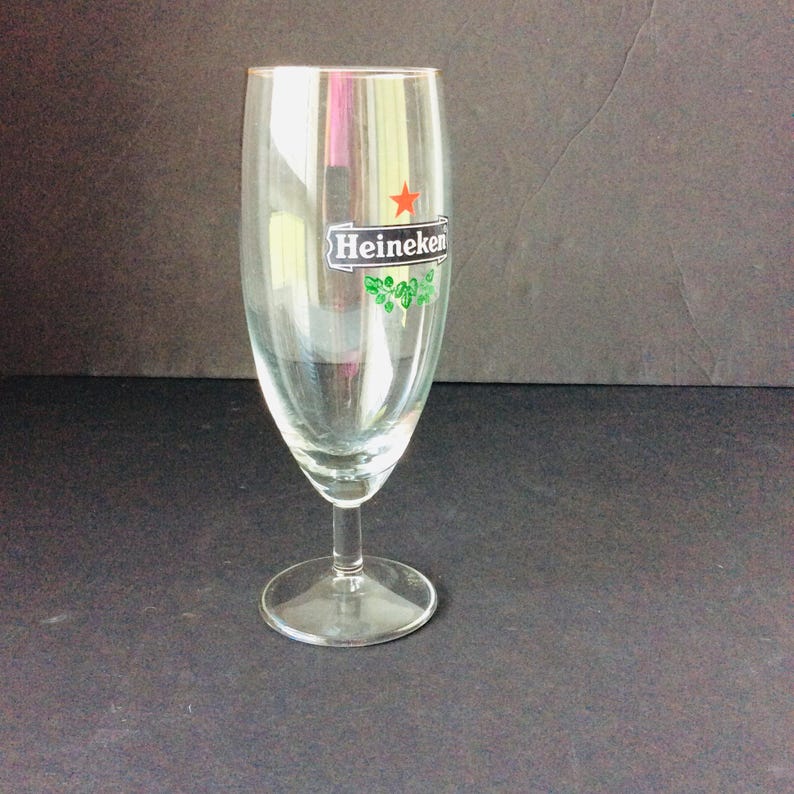 May include: A clear, stemmed beer glass with a wide bowl and a narrow base. The glass features the Heineken logo, including a red star and green accents. The glass is empty and sits on a dark surface.
