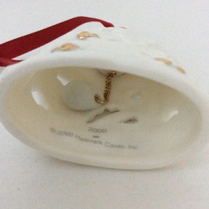 May include: Cream-colored bell ornament with gold accents and a red ribbon. The bell has a gold chain and a small white ball inside. The text "2000 Hallmark Cards, Inc." is printed on the bell.