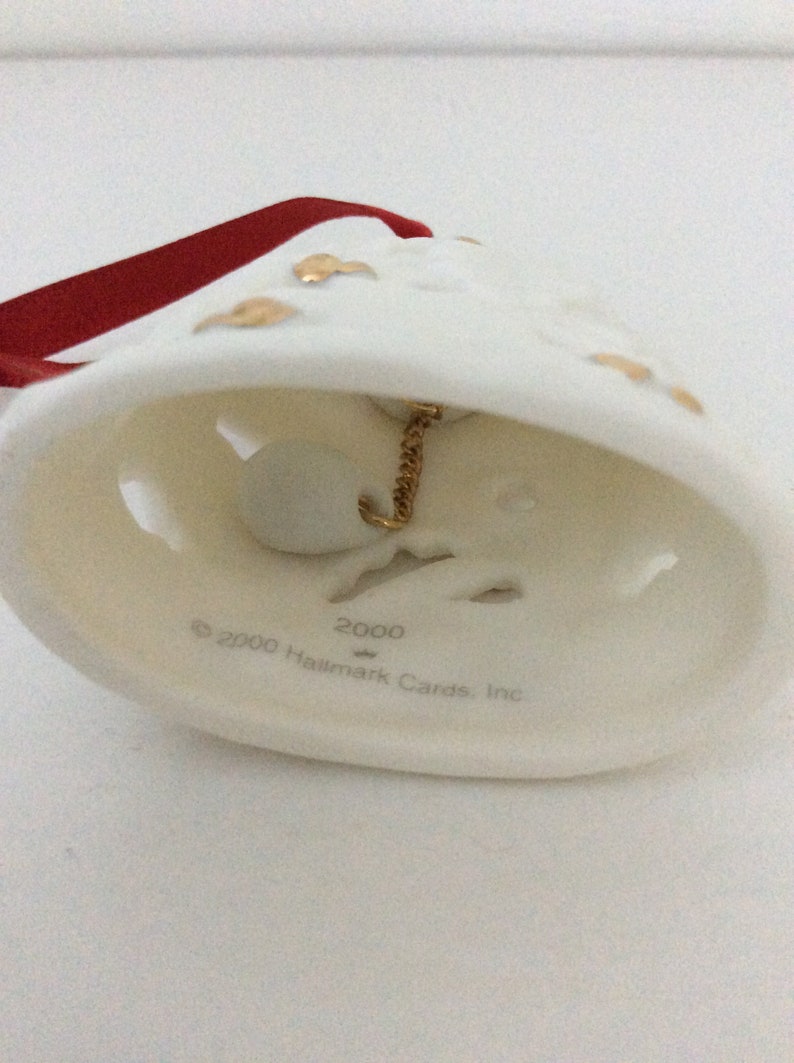 May include: Cream-colored bell ornament with gold accents and a red ribbon. The bell has a gold chain and a small white ball inside. The text "2000 Hallmark Cards, Inc." is printed on the bell.