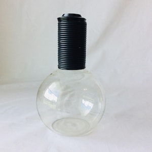 May include: A clear glass vase with a round base and a cylindrical black top. The black top has a ribbed texture. The vase is empty and sits on a white surface.