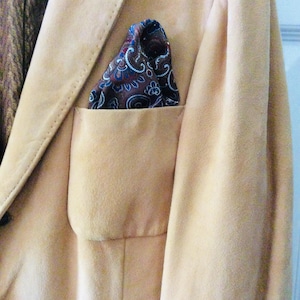 May include: A light tan blazer featuring a breast pocket with a folded pocket square. The pocket square is dark brown with a detailed paisley pattern in shades of blue, white, and red. The blazer has visible stitching along the lapel edge.