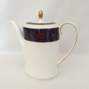 May include: A white porcelain teapot featuring a cylindrical body with a gold-trimmed handle and spout. The upper section is decorated with a dark blue and orange paisley pattern, finished with a gold-toned finial on the lid.