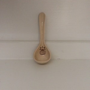 May include: A small, light beige ceramic spoon with a speckled texture. The bowl of the spoon features a dark brown line drawing of a stylized pumpkin or fruit with a stem and two small leaves centered on the interior surface of the spoon.