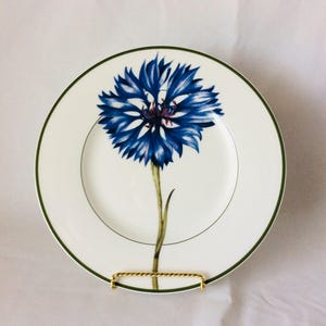 May include: A white porcelain plate with a green rim features a blue and purple flower with a green stem. The plate is on a gold stand.