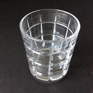 Op de afbeelding: Een helder glazen drinkglas met een rasterpatroon van verhoogde vierkanten op het buitenoppervlak. De beker heeft een brede, ronde rand en een licht taps toelopende cilindrische vorm, wat zorgt voor een transparant ontwerp.
