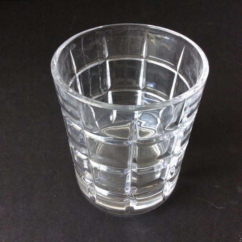 Op de afbeelding: Een helder glazen drinkglas met een rasterpatroon van verhoogde vierkanten op het buitenoppervlak. De beker heeft een brede, ronde rand en een licht taps toelopende cilindrische vorm, wat zorgt voor een transparant ontwerp.
