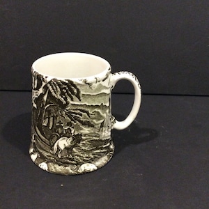 May include: A ceramic mug featuring a black and white transferware print of a landscape scene with figures on horseback, trees, and rolling hills. The mug has a cylindrical body, a wide base, and a curved handle, with a white interior finish.