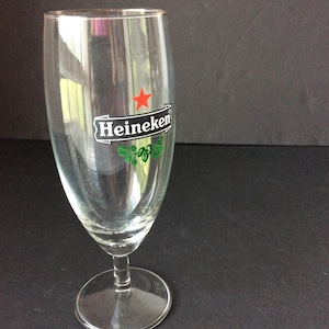 May include: A clear, stemmed beer glass with the Heineken logo. The logo features a red star, the word "Heineken" in white, and green leaf accents. The glass has a wide bowl and a round base.