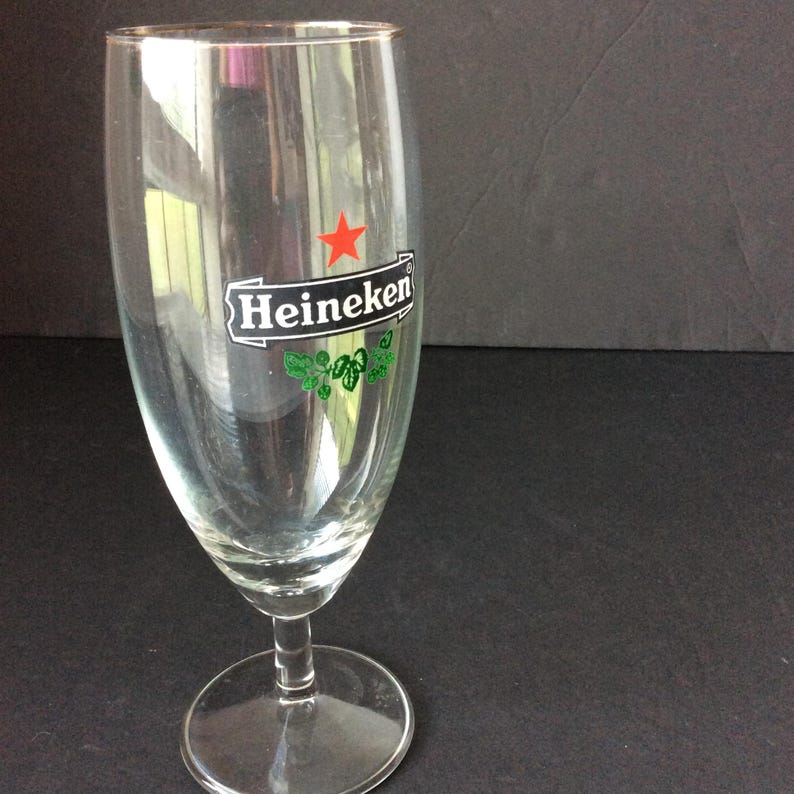 May include: A clear, stemmed beer glass with the Heineken logo. The logo features a red star, the word "Heineken" in white, and green leaf accents. The glass has a wide bowl and a round base.