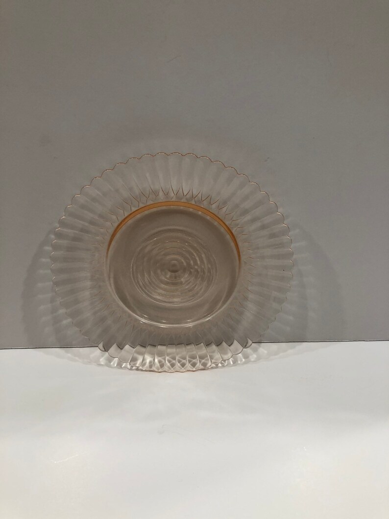 Puede incluir: Un plato de cristal transparente te&ntilde;ido de rosa, con borde festoneado. El plato tiene un dise&ntilde;o acanalado que irradia desde el centro, con un borde dorado. El plato es probablemente vintage o antiguo, adecuado para servir o exhibir.