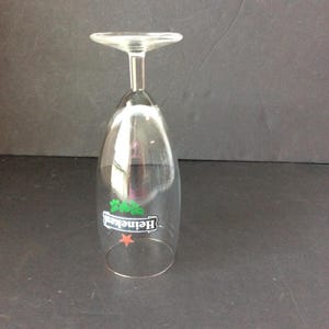 May include: Clear, inverted Heineken beer glass with a stem and a wide, flat base. The glass features the Heineken logo in green and white with a red star. The glass is made of clear glass.