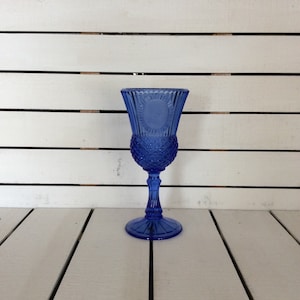 May include: A vibrant cobalt blue glass goblet featuring a textured diamond pattern on the lower bowl, vertical fluting on the upper rim, and a circular medallion detail. The stem is faceted and leads to a wide, circular, flat base.