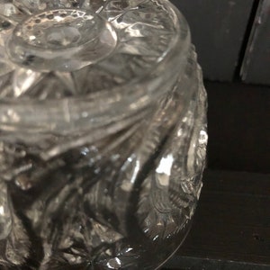 May include: Close-up of a clear, cut-glass jar with a detailed, textured pattern. The jar has a rounded shape and a decorative lid. The glass reflects light, highlighting the intricate design. The jar sits on a dark surface.