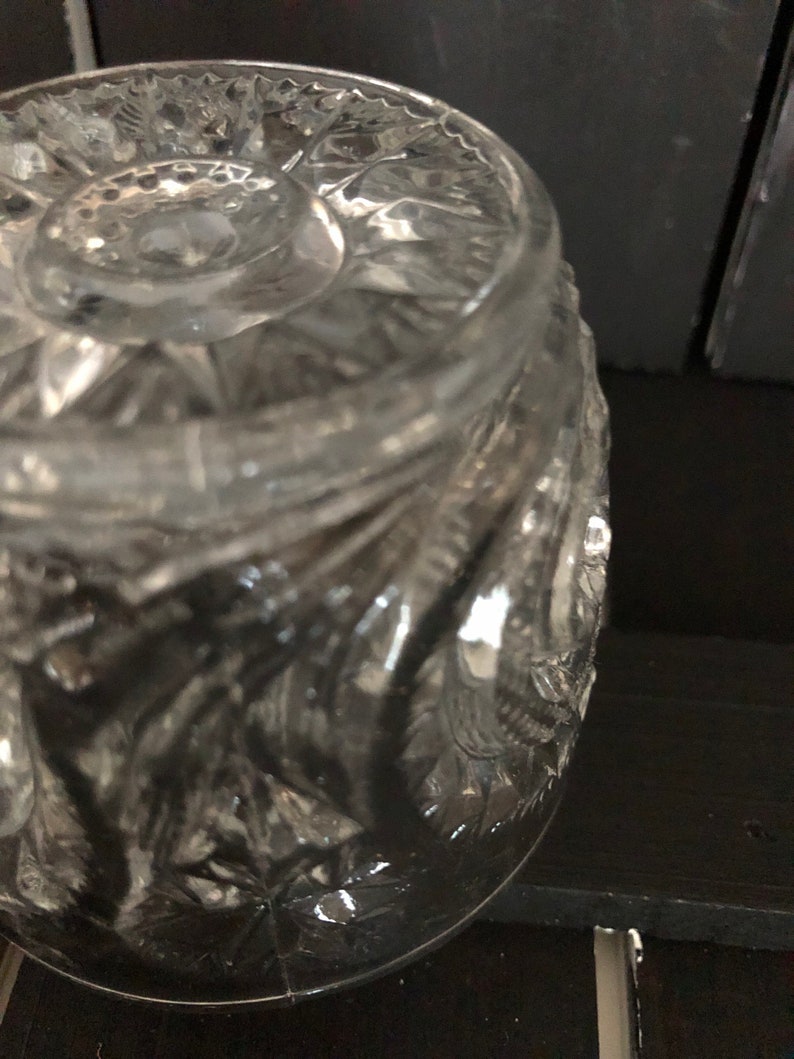 May include: Close-up of a clear, cut-glass jar with a detailed, textured pattern. The jar has a rounded shape and a decorative lid. The glass reflects light, highlighting the intricate design. The jar sits on a dark surface.